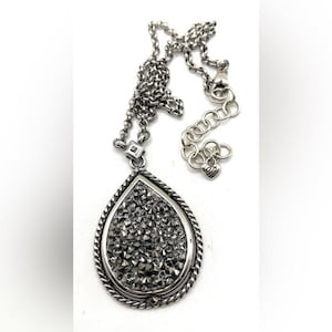 May include: A silver-toned necklace with a teardrop pendant. The pendant is filled with small, sparkling gray stones and framed by a twisted rope design. The chain is also silver and has an adjustable length.