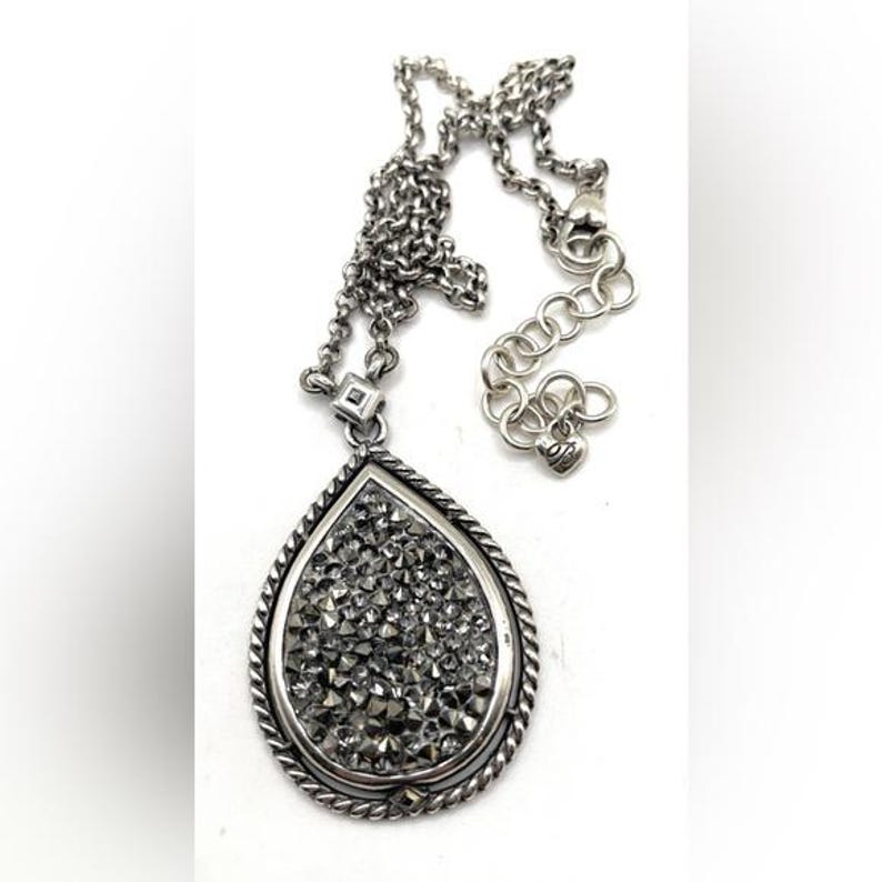 May include: A silver-toned necklace with a teardrop pendant. The pendant is filled with small, sparkling gray stones and framed by a twisted rope design. The chain is also silver and has an adjustable length.