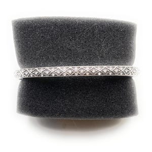 May include: A silver bangle bracelet with a repeating pattern of diamond-shaped designs, each containing a small, clear gemstone. The bracelet is displayed on a dark gray, textured surface against a white background.