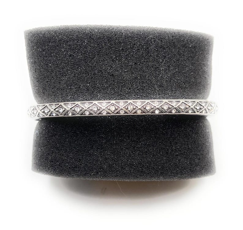 May include: A silver bangle bracelet with a repeating pattern of diamond-shaped designs, each containing a small, clear gemstone. The bracelet is displayed on a dark gray, textured surface against a white background.