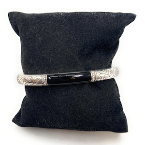 May include: A silver bangle bracelet with a black enamel inlay and a swirl design. The bracelet is on a black velvet cushion.