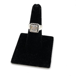 May include: A silver ring with a square, clear gemstone. The gemstone is set within a square frame with a rope-like design. The ring is displayed on a black ring holder against a white background.