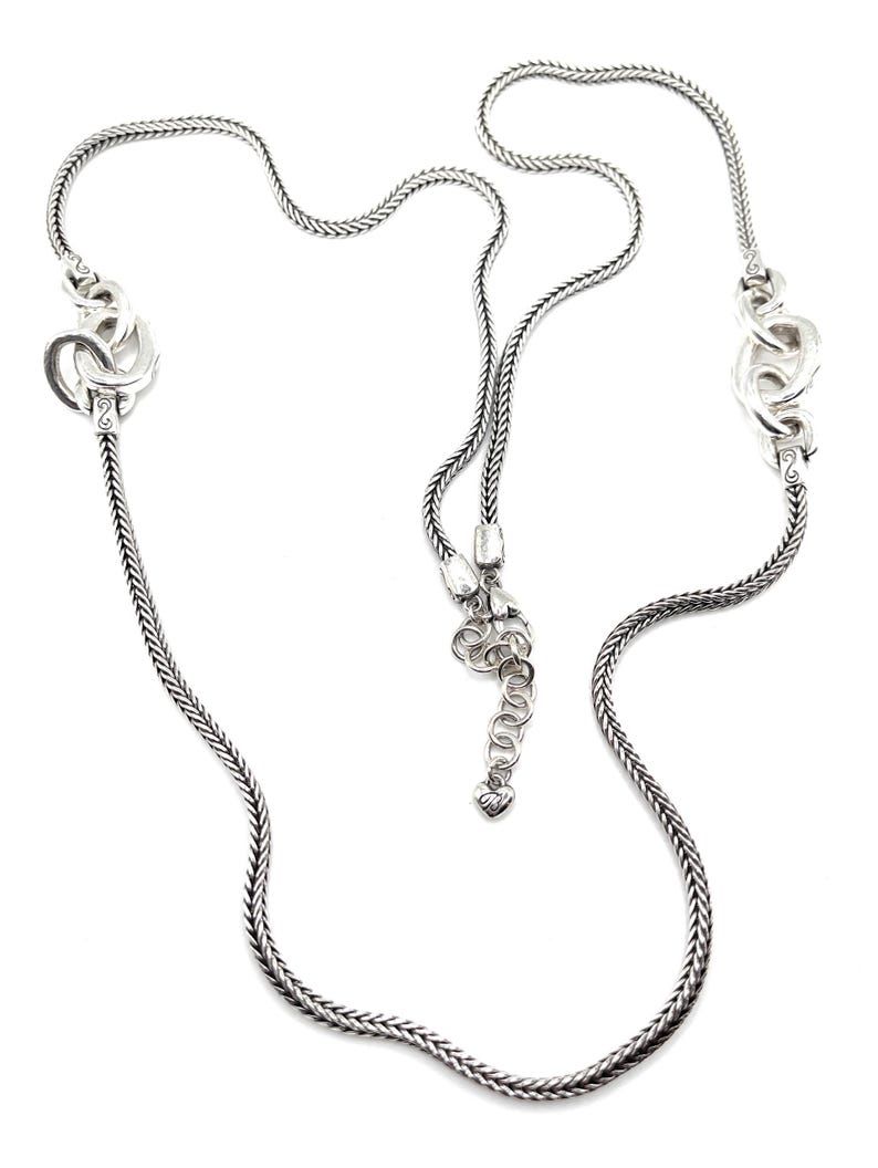 Puede incluir: Un collar plateado con un dise&ntilde;o de cadena trenzada. El collar presenta eslabones decorativos y estilizados a intervalos a lo largo de la cadena. El collar tiene una longitud ajustable con un peque&ntilde;o dije en forma de coraz&oacute;n al final.