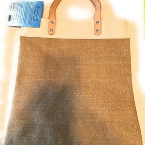 Puede incluir: Una bolsa de lona beige con asas de cuero marr&oacute;n y un fondo de arpillera. La bolsa tiene una etiqueta blanca con el texto "You make the Earth go round!" en ella.
