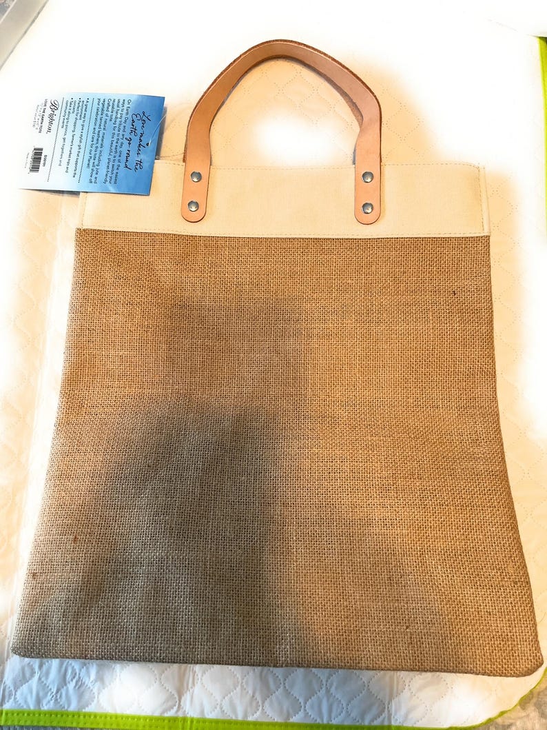 Puede incluir: Una bolsa de lona beige con asas de cuero marr&oacute;n y un fondo de arpillera. La bolsa tiene una etiqueta blanca con el texto "You make the Earth go round!" en ella.