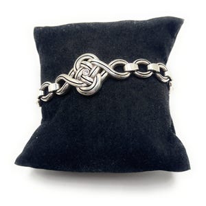 May include: A silver-toned bracelet with a Celtic knot design and a small, clear gemstone. The bracelet is displayed on a black velvet pillow against a white background. The bracelet features a series of interlocking links.