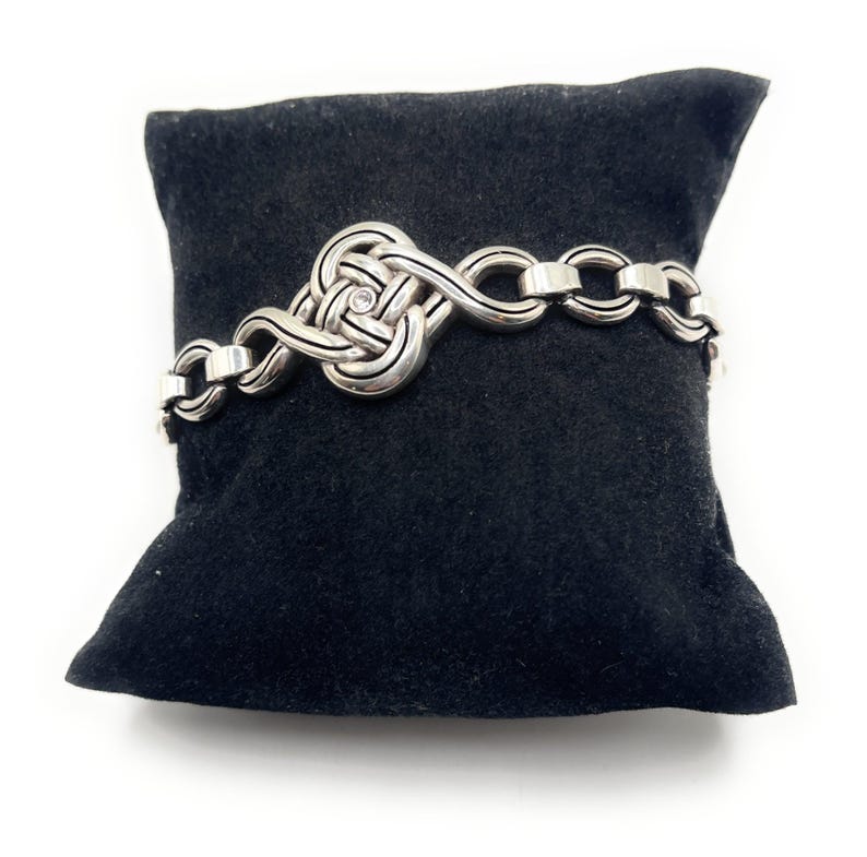 May include: A silver-toned bracelet with a Celtic knot design and a small, clear gemstone. The bracelet is displayed on a black velvet pillow against a white background. The bracelet features a series of interlocking links.