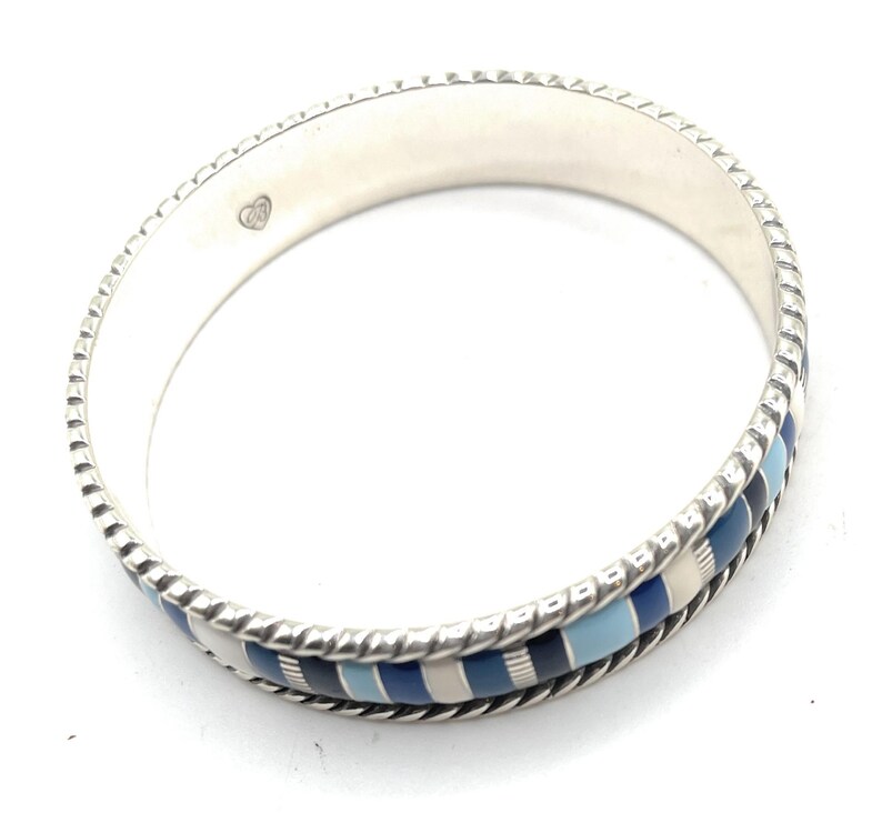 Może przedstawiać: Srebrna bransoletka obręczowa z teksturowaną krawędzią i wzorem niebieskich, jasnoniebieskich i białych prostokątnych segment&oacute;w. Bransoletka ma logo w kształcie serca po wewnętrznej stronie. Stylowa biżuteria.