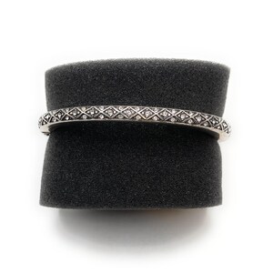 May include: Silver-toned bracelet with a repeating diamond pattern, each diamond set with small, clear crystals. The bracelet is displayed on a black, textured surface against a white background. The jewelry piece is a bangle style.