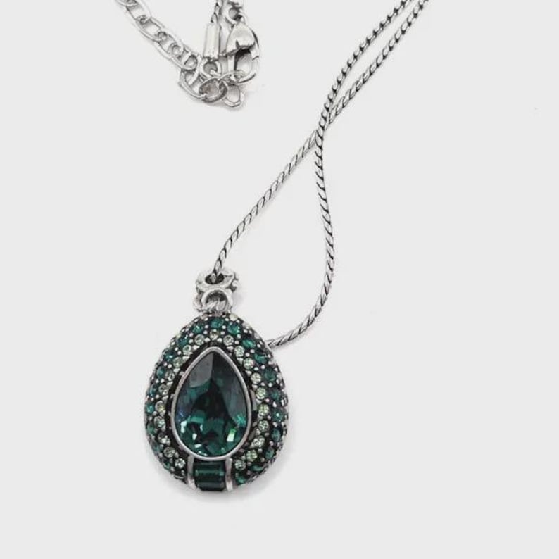 K&ouml;nnte beinhalten: Silberfarbene Halskette mit einem Anh&auml;nger. Der Anh&auml;nger hat einen gro&szlig;en, tropfenf&ouml;rmigen, smaragdgr&uuml;nen Edelstein, umgeben von kleineren gr&uuml;nen Steinen. Die Kette ist filigran und der Verschluss ist sichtbar.