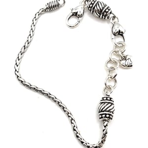 May include: A silver chain bracelet with a braided chain, a heart-shaped clasp, and two decorative beads.