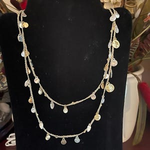 May include: A double-strand necklace featuring a light-colored braided cord adorned with numerous small, circular metallic charms in alternating silver and gold tones, spaced evenly along the length of the two draped strands.