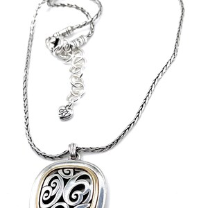 May include: Silver-tone necklace with a square pendant featuring a gold-tone border and an openwork design with swirling patterns. The necklace has an adjustable chain and a heart-shaped charm.