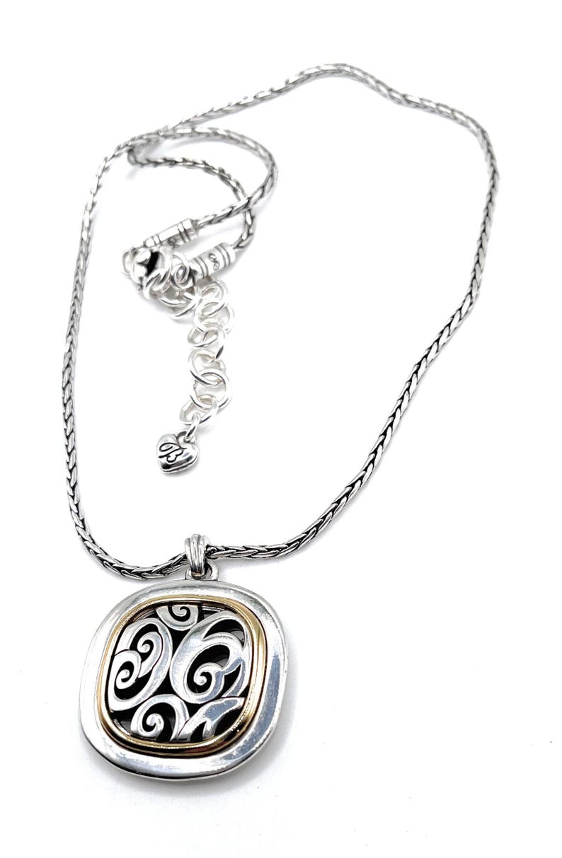 May include: Silver-tone necklace with a square pendant featuring a gold-tone border and an openwork design with swirling patterns. The necklace has an adjustable chain and a heart-shaped charm.