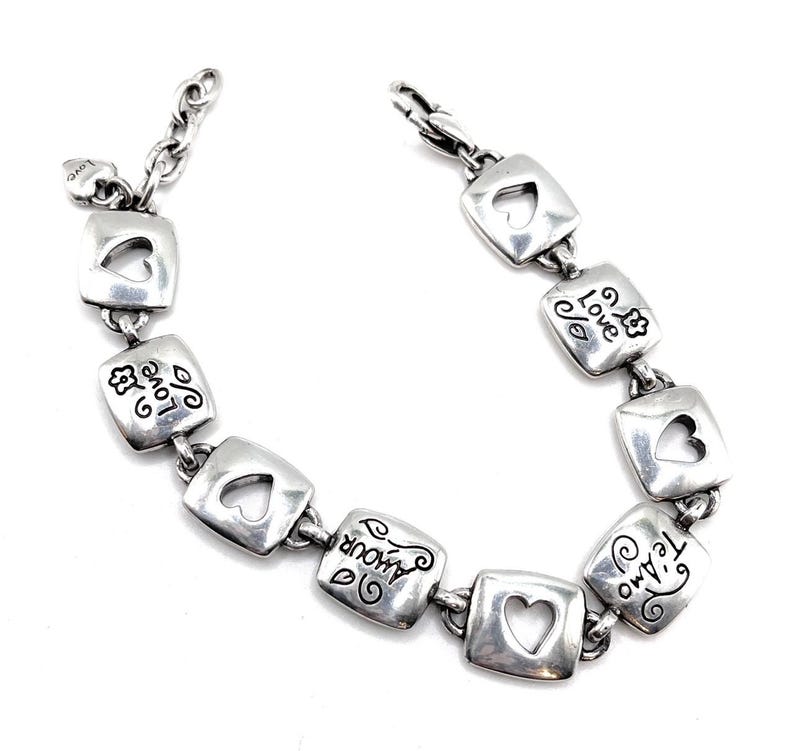 May include: Silver bracelet with square links, each with a heart-shaped cutout. The links are engraved with words such as "love" and "amour." It has a lobster clasp and a small heart charm.