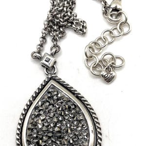 May include: Silver-toned necklace with a teardrop pendant. The pendant is filled with small, sparkling, gray crystals and framed by a twisted rope design. The chain has a lobster clasp and an adjustable length.
