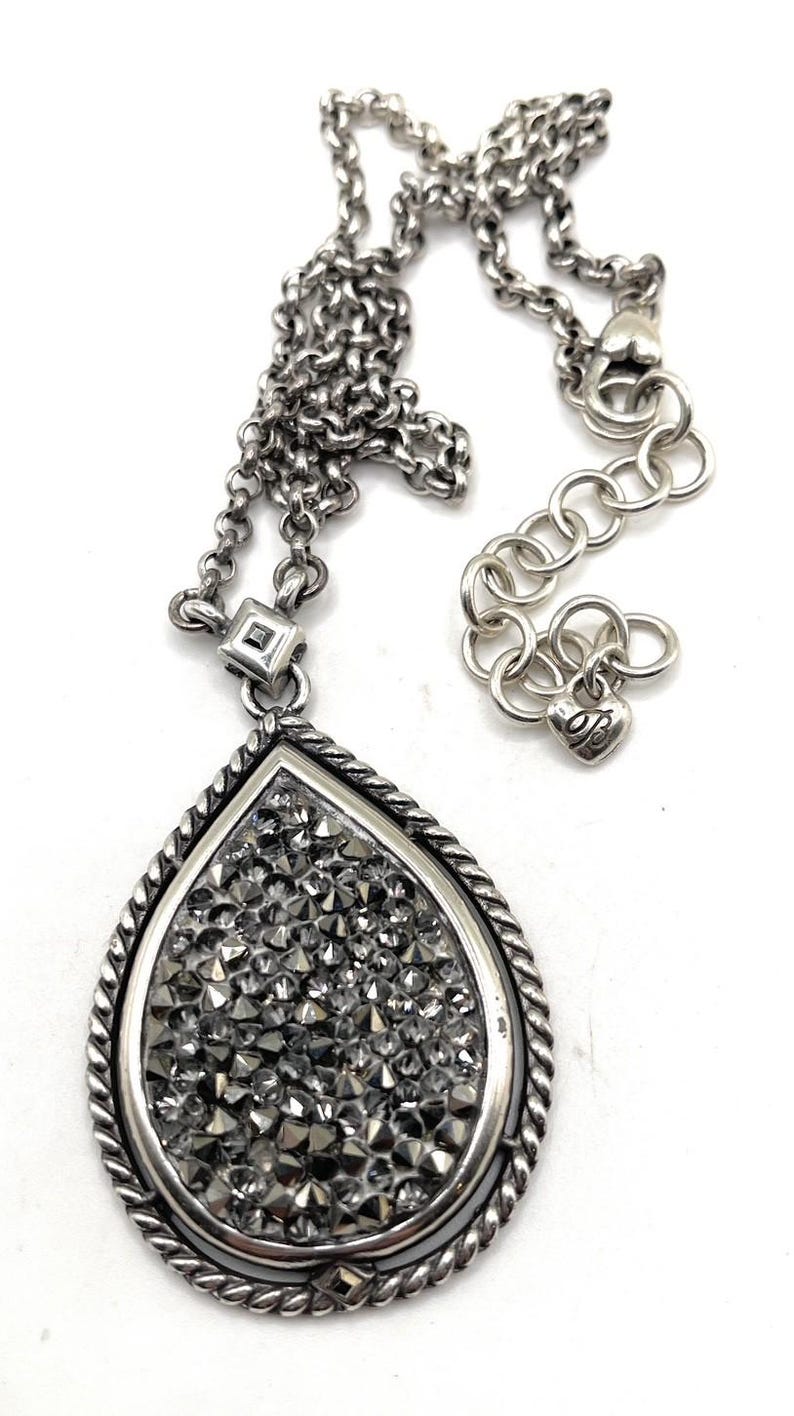 May include: Silver-toned necklace with a teardrop pendant. The pendant is filled with small, sparkling, gray crystals and framed by a twisted rope design. The chain has a lobster clasp and an adjustable length.