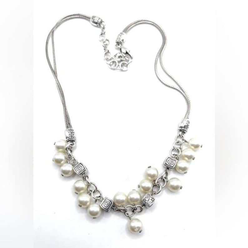 May include: Silver-toned necklace featuring a delicate chain and a cluster of faux pearls. The necklace has decorative square links with a swirl design. The necklace is a classic piece of jewelry.