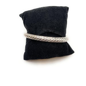 May include: A silver-coloured bangle bracelet with a twisted rope design. The bracelet is presented on a black velvet cushion against a white backdrop. The bangle is a solid, circular band with a textured surface.