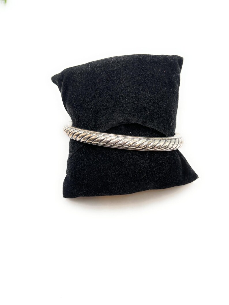 May include: A silver-coloured bangle bracelet with a twisted rope design. The bracelet is presented on a black velvet cushion against a white backdrop. The bangle is a solid, circular band with a textured surface.