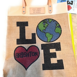 Puede incluir: Una bolsa de lona beige con asas de cuero marr&oacute;n. La bolsa tiene un gran gr&aacute;fico de la palabra "LOVE" con un coraz&oacute;n rojo que dice "BRIGHTON" en el medio de la "O" y un globo terr&aacute;queo azul y verde en el medio de la "V". La bolsa tambi&eacute;n tiene una peque&ntilde;a etiqueta blanca que dice "LOVE MAKES THE EARTH GO ROUND".