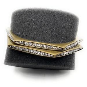 May include: Two gold-toned, geometric bangles with a row of clear crystals. The bracelets are stacked on a dark gray foam cylinder. The jewelry has a modern, minimalist design.