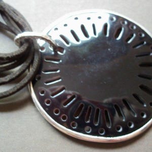 Könnte beinhalten: Eine schwarze Halskette mit einem silbernen runden Anhänger mit einem ausgeschnittenen Design. Der Anhänger hat eine glänzende, reflektierende Oberfläche.