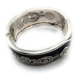 May include: Silver-tone hinged bracelet with a black enamel band. The bracelet features intricate scrollwork and sparkling crystal accents. The interior has decorative etched patterns. A stylish piece of jewelry.