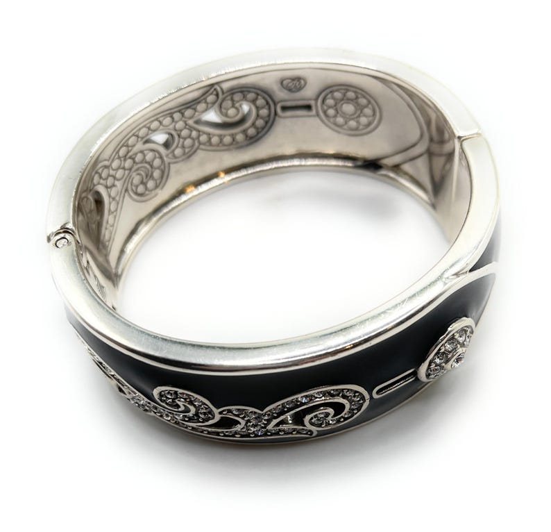 May include: Silver-tone hinged bracelet with a black enamel band. The bracelet features intricate scrollwork and sparkling crystal accents. The interior has decorative etched patterns. A stylish piece of jewelry.
