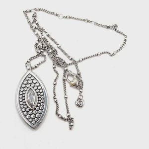 Puede incluir: Collar plateado con un colgante ovalado grande. El colgante presenta un cristal transparente central rodeado de peque&ntilde;os puntos plateados en relieve. La cadena es una mezcla de secciones enlazadas y con cuentas, con un cierre en forma de coraz&oacute;n y un peque&ntilde;o dije de coraz&oacute;n.