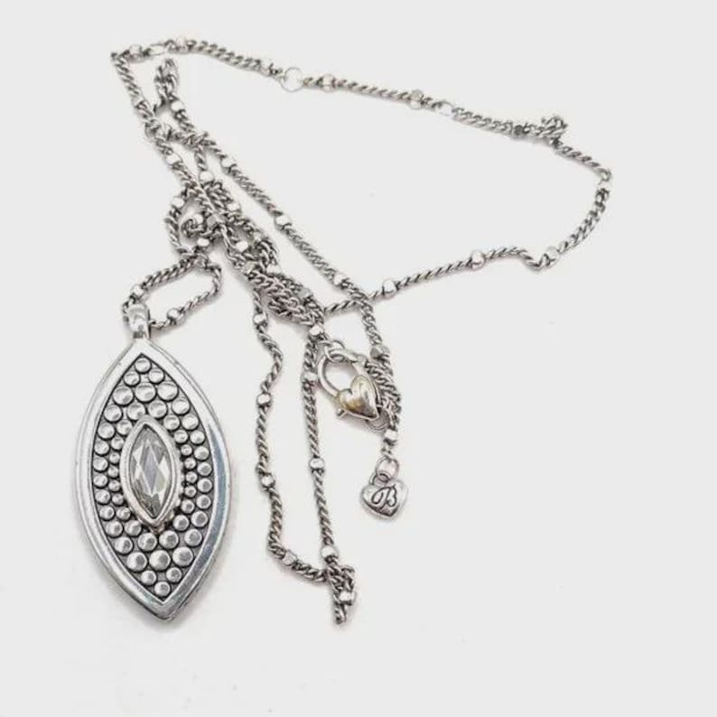 Puede incluir: Collar plateado con un colgante ovalado grande. El colgante presenta un cristal transparente central rodeado de peque&ntilde;os puntos plateados en relieve. La cadena es una mezcla de secciones enlazadas y con cuentas, con un cierre en forma de coraz&oacute;n y un peque&ntilde;o dije de coraz&oacute;n.