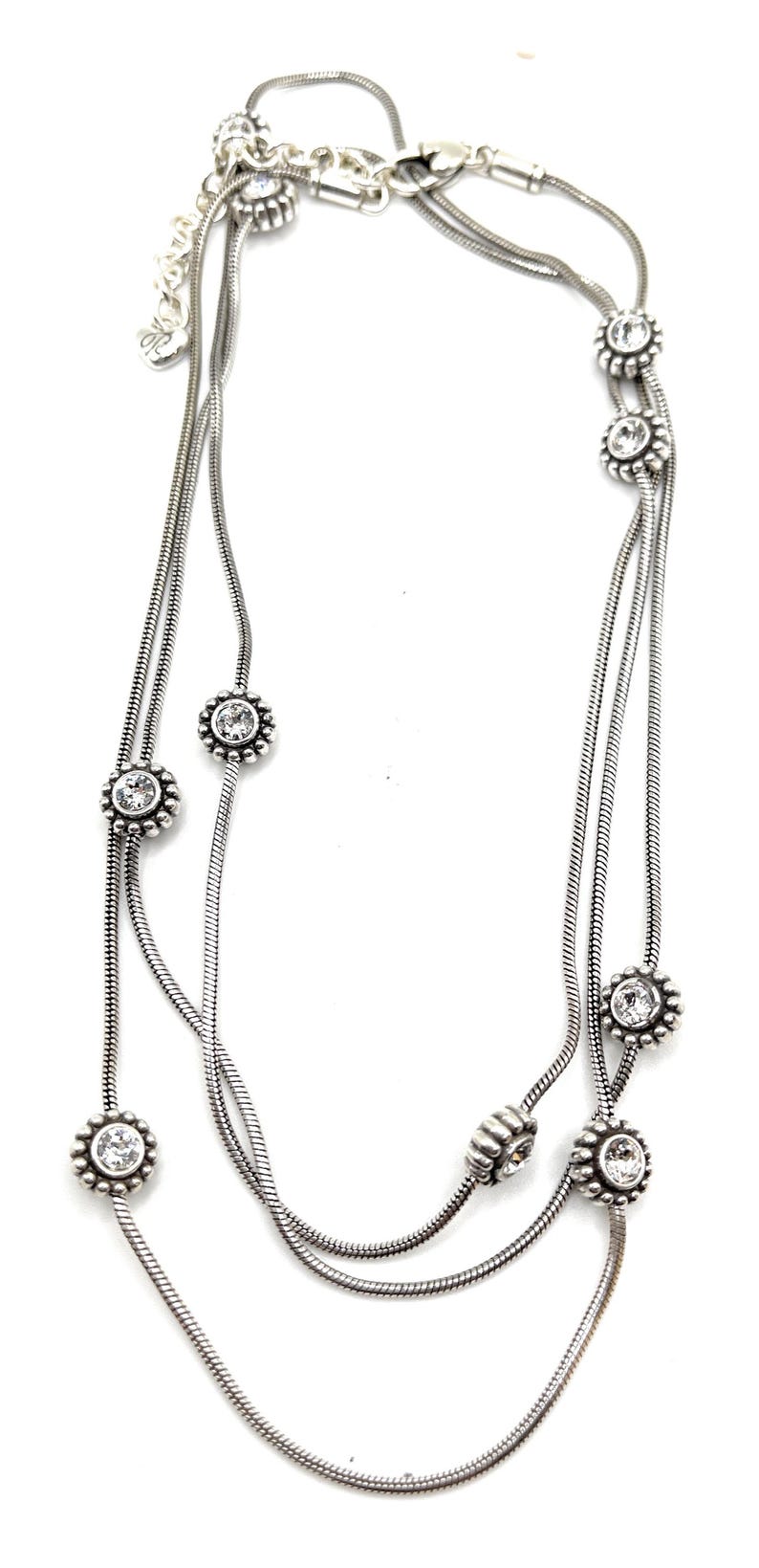 Może przedstawiać: Srebrny naszyjnik z trzema sznurami. Każdy sznur ma przezroczyste, okrągłe kamienie szlachetne osadzone w ramie w kształcie kwiatu. Naszyjnik ma delikatny, elegancki design, odpowiedni na r&oacute;żne okazje.