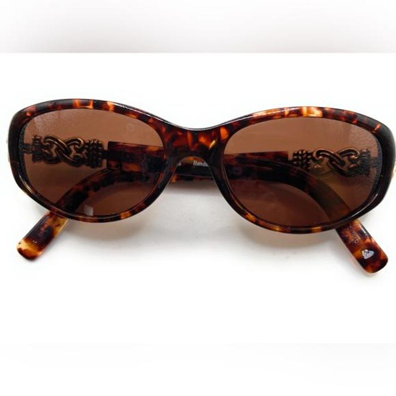 Puede incluir: Gafas de sol ovaladas con montura con estampado de carey y lentes marrones. La montura presenta detalles decorativos en forma de cadena en las patillas. Las gafas de sol est&aacute;n sobre un fondo blanco.