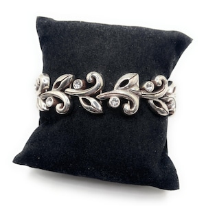 Puede incluir: Pulsera plateada con dise&ntilde;o de hojas y espirales, adornada con gemas redondas y transparentes. La pulsera se exhibe sobre una superficie de terciopelo negro, mostrando sus intrincados detalles y su estilo elegante.