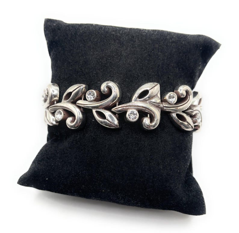 Puede incluir: Pulsera plateada con dise&ntilde;o de hojas y espirales, adornada con gemas redondas y transparentes. La pulsera se exhibe sobre una superficie de terciopelo negro, mostrando sus intrincados detalles y su estilo elegante.