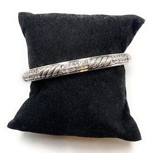 May include: A silver bangle bracelet with a twisted rope pattern and a line of clear crystals. The bracelet is presented on a black velvet cushion against a white backdrop. A classic piece of jewellery.