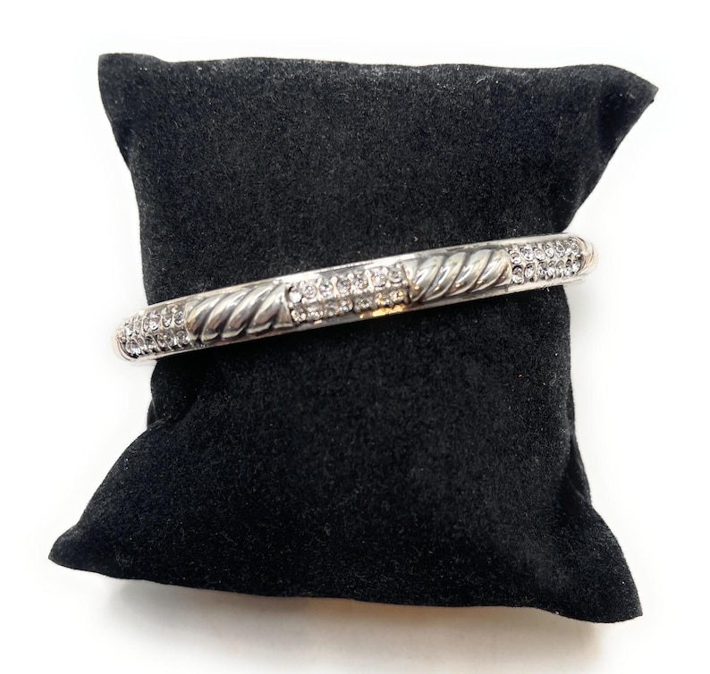 May include: A silver bangle bracelet with a twisted rope pattern and a line of clear crystals. The bracelet is presented on a black velvet cushion against a white backdrop. A classic piece of jewellery.