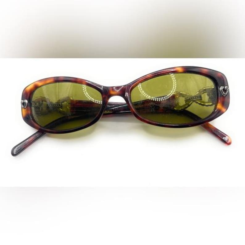 Puede incluir: Gafas de sol con montura ovalada de carey y lentes tintadas en verde. Las monturas marrones presentan un dise&ntilde;o de cadena en las patillas. Las gafas de sol est&aacute;n sobre un fondo blanco.