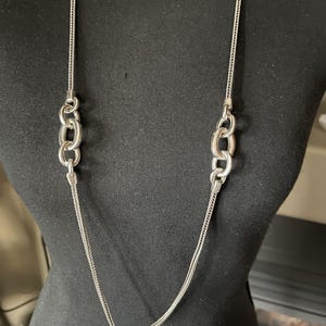 Puede incluir: Un collar plateado con una cadena larga y delicada y dos conjuntos de tres eslabones ovalados entrelazados. El collar se exhibe en un maniqu&iacute; negro, mostrando su longitud y dise&ntilde;o. La cadena tiene una textura tejida.