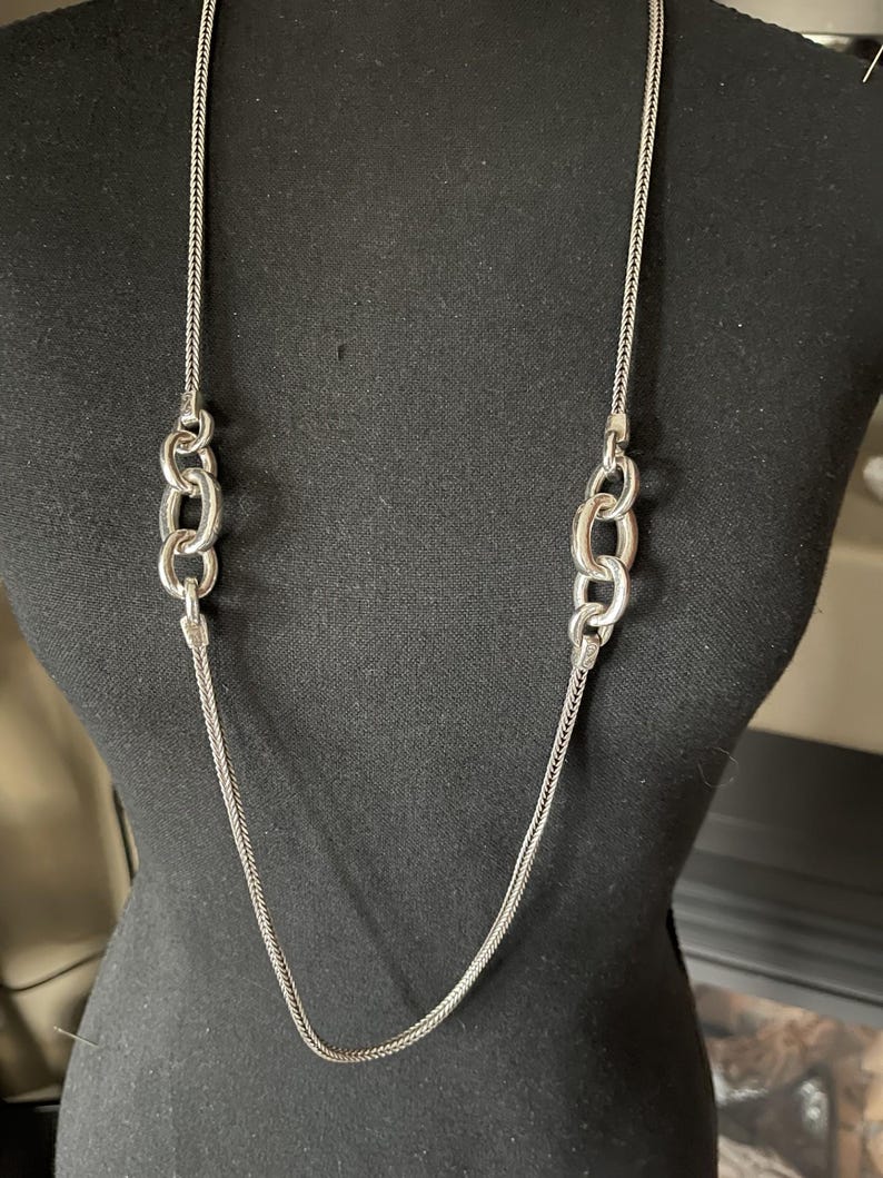 Puede incluir: Un collar plateado con una cadena larga y delicada y dos conjuntos de tres eslabones ovalados entrelazados. El collar se exhibe en un maniqu&iacute; negro, mostrando su longitud y dise&ntilde;o. La cadena tiene una textura tejida.