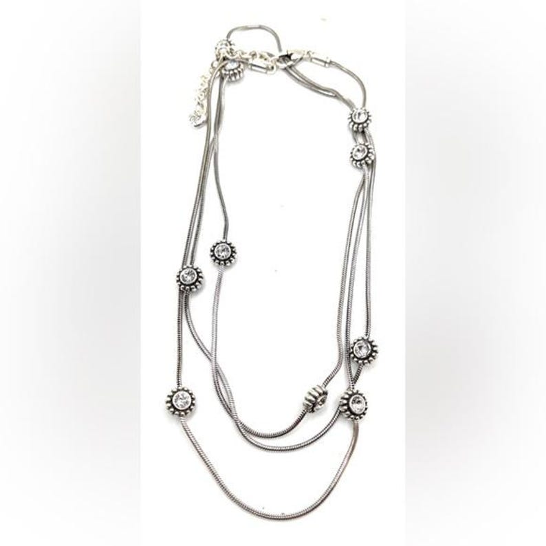Może przedstawiać: Srebrny naszyjnik z trzema sznurami. Każdy sznur ma przezroczyste, okrągłe kamienie szlachetne w kwiatowym wzorze. Naszyjnik ma zapięcie na karabińczyk.