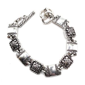 May include: Silver bracelet with a toggle clasp and ornate rectangular links. Each link features a floral design.