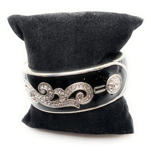 May include: A black enamel hinged bangle bracelet with silver-tone trim and a decorative swirl design encrusted with clear crystals. The bracelet features a round crystal accent and is displayed on a black velvet pillow.