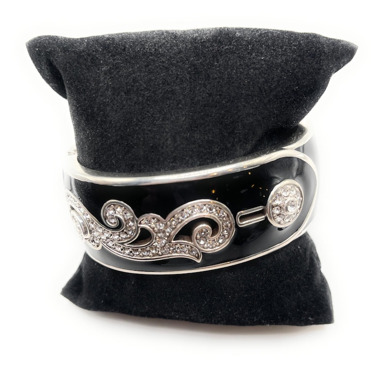 May include: A black enamel hinged bangle bracelet with silver-tone trim and a decorative swirl design encrusted with clear crystals. The bracelet features a round crystal accent and is displayed on a black velvet pillow.