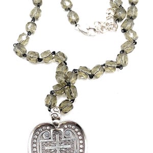 Peut inclure: Collier argent&eacute; avec un pendentif en forme de c&oelig;ur orn&eacute; d'une croix. Le pendentif pr&eacute;sente des d&eacute;tails de volutes et une bordure perl&eacute;e. Le collier est compos&eacute; de perles facett&eacute;es gris fum&eacute; et d'un fermoir argent&eacute;. Longueur d'environ 45 cm.