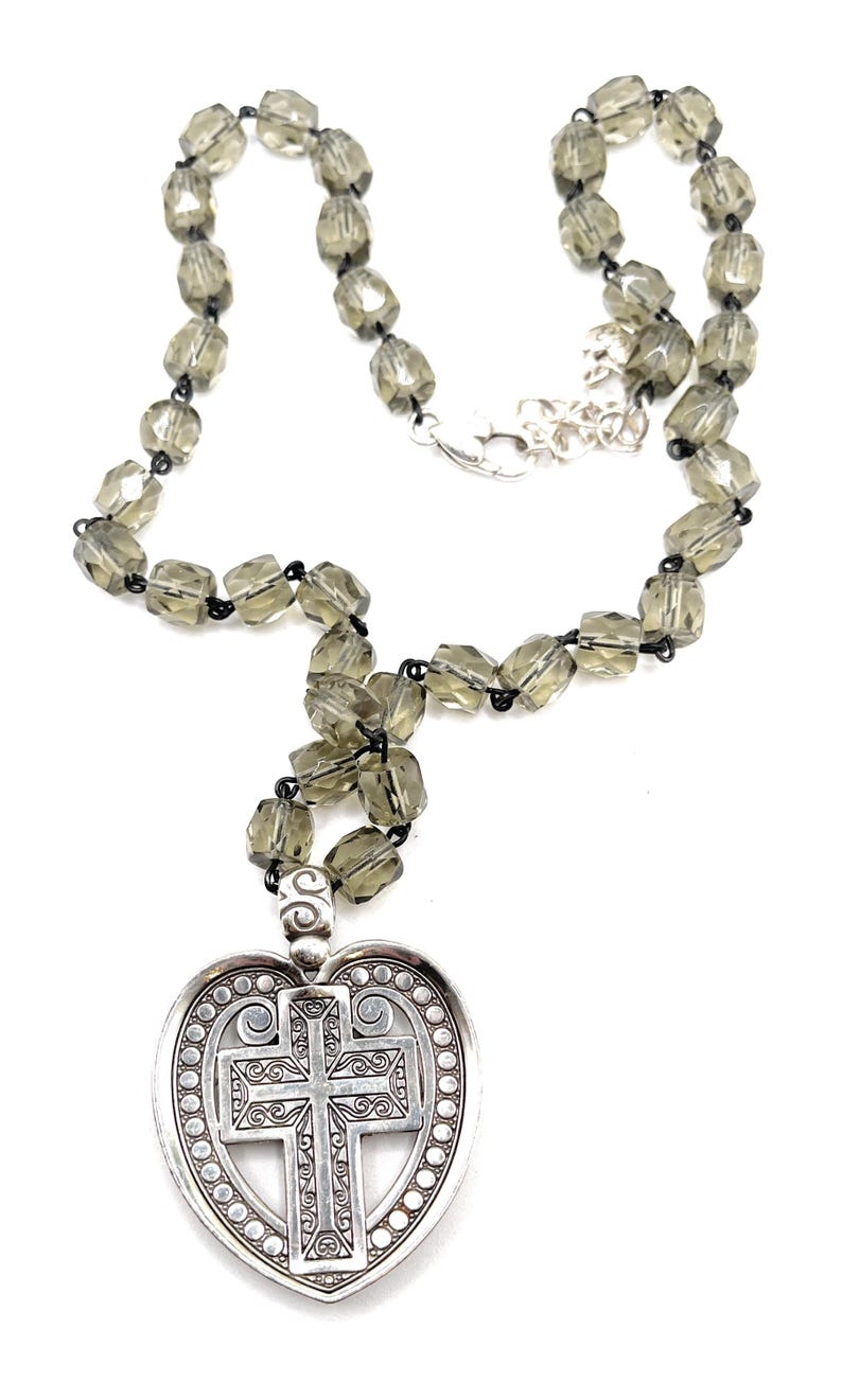 Peut inclure: Collier argent&eacute; avec un pendentif en forme de c&oelig;ur orn&eacute; d'une croix. Le pendentif pr&eacute;sente des d&eacute;tails de volutes et une bordure perl&eacute;e. Le collier est compos&eacute; de perles facett&eacute;es gris fum&eacute; et d'un fermoir argent&eacute;. Longueur d'environ 45 cm.