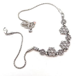 May include: A silver-toned necklace featuring a series of stylized flower-shaped charms. The necklace has a delicate chain and a lobster clasp closure. The design includes a small charm with a heart and a crystal accent.