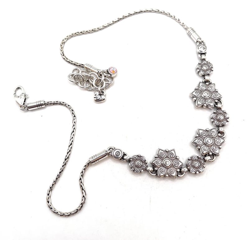 May include: A silver-toned necklace featuring a series of stylized flower-shaped charms. The necklace has a delicate chain and a lobster clasp closure. The design includes a small charm with a heart and a crystal accent.