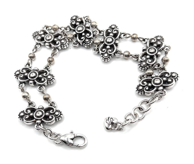 May include: Silver-toned bracelet featuring ornate floral and scrollwork designs. The bracelet has a series of decorative links connected by small, round beads. It includes a lobster clasp closure and an adjustable chain.