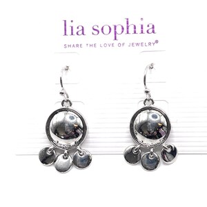 Puede incluir: Pendientes colgantes plateados. Cada pendiente presenta un colgante circular grande y reflectante con tres dijes circulares más pequeños colgando debajo. Los pendientes se exhiben en una tarjeta blanca con el texto "lia sophia" y "SHARE THE LOVE OF JEWELRY".